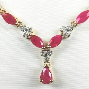 May include: A gold necklace featuring marquise-cut red gemstones and a central pear-shaped red gemstone drop. The design includes small clusters of clear, round-cut stones set in silver-toned metal accents between the red marquise-cut stones.