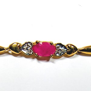 May include: A gold-tone bracelet featuring a central oval-cut vibrant pink gemstone held in a four-prong setting. The centre stone is flanked by two heart-shaped accents, each containing small clear stones, followed by smooth gold-tone metal links.