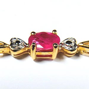 May include: A gold bracelet featuring a central oval-cut vibrant red gemstone held by four prongs. Flanking the centre stone are two small heart-shaped accents, each set with a tiny clear stone, connected to a polished gold chain link band.