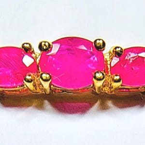 May include: A gold-toned bracelet featuring three vibrant, oval-cut hot pink stones set in prong settings. The central stone is slightly larger than the two flanking stones, and the metal band consists of smooth, polished gold-coloured links.