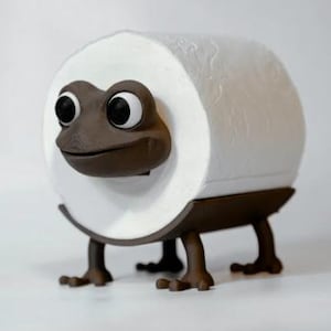 May include: A whimsical brown frog-shaped toilet paper holder. The holder features a roll of white toilet paper, with the frog's face peeking through. The frog has large, expressive eyes and is supported by four brown legs.
