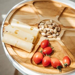 Peut inclure: Un plateau de service rond en bois avec plusieurs compartiments. Il contient des tranches de fromage roulées, des tomates cerises rouges et des pistaches. Le plateau est posé sur une table blanche.