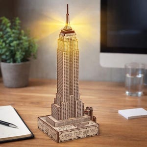 Könnte beinhalten: Ein detailliertes Holzmodell des Empire State Building mit warmgelben LED-Leuchten. Das braune Modell steht auf einem Sockel mit der Aufschrift "Empire State Building" und "USA 1930-1931". Das Modell steht auf einem Holztisch.