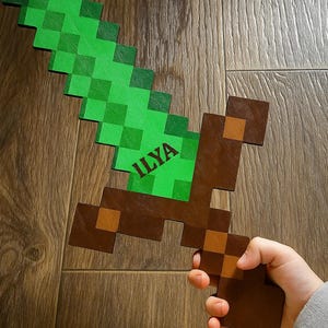 May include: A pixelated green and brown sword toy, held by a hand. The sword has a green blade and a brown handle. The word "ILYA" is printed on the blade. The toy is made of wood and is designed to look like a sword from a video game.