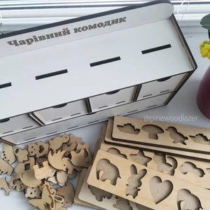 Puede incluir: Un organizador de madera blanco con cajones y el texto "Чарівний комодник" en gris. También hay piezas de rompecabezas de madera con forma de animales y tableros de rompecabezas con formas recortadas. El organizador está diseñado para guardar objetos pequeños.