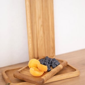Könnte beinhalten: Serviertabletts aus Holz in verschiedenen Größen. Ein Tablett steht aufrecht, während zwei weitere gestapelt sind. Die Tabletts sind mit frischen Aprikosen und Blaubeeren gefüllt. Das Holz hat eine natürliche Maserung und eine warme, hellbraune Farbe.