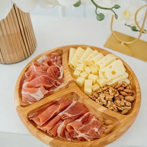 May include: A wooden serving platter with three compartments, filled with sliced prosciutto, various cheeses, and almonds. The platter is made of light-colored wood and is arranged on a white surface. The food is ready to serve.