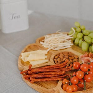 Peut inclure: Un plateau de service en bois avec plusieurs compartiments, rempli d'une variété de collations. Le plateau contient du fromage, des amandes, des raisins, des tomates cerises et des bâtonnets de viande. Un récipient à thé blanc est en arrière-plan.