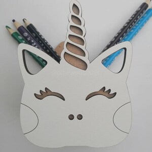 Op de afbeelding: Een witte houten eenhoornvormige potloodhouder met een bruine hoorn en oogdetails. De houder bevat verschillende kleurpotloden. Het ontwerp omvat een uitgesneden gezicht en oren.