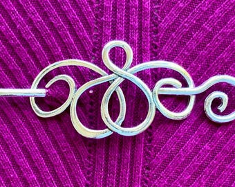 May include: A silver metal shawl pin with a decorative, intricate design. The pin has a long, thin shaft and a decorative, swirling design at the top.