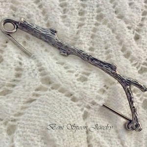 May include: A silver twig-shaped brooch with a textured surface. The brooch is designed to look like a small branch with a single fork. The pin is attached to the back of the brooch.