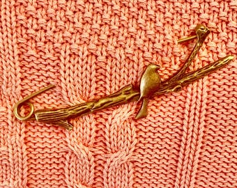 May include: A gold-toned metal brooch featuring two birds perched on a branch. The brooch is designed with a safety pin closure and is set against a pink knitted background.