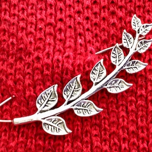 May include: A silver-toned leaf branch brooch with a safety pin closure. The brooch is on a red knitted fabric background.