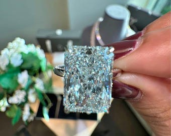 Può includere: Un anello con diamante taglio radiant. Il grande diamante rettangolare è il punto focale, con una superficie brillante e sfaccettata. La fascia dell'anello è color argento e il diamante è incastonato con pietre più piccole. L'anello è tenuto da una mano.