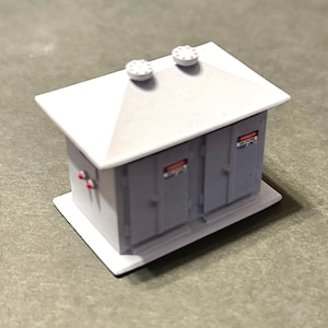 May include: A miniature gray utility building model with a white roof and base. The model has two doors, two circular vents, and red warning labels. The building is a detailed replica, likely for a model train set or architectural display.