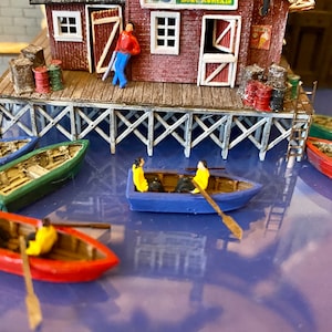 May include: Miniature model of a waterfront scene with a red building, pier, and several small boats. The boats are painted in various colors, including blue, green, and red. Figures are in some of the boats and on the pier.