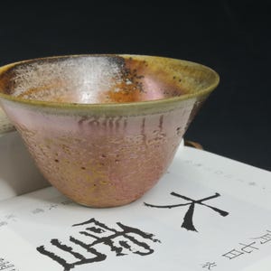 May include: A handmade ceramic bowl with a pink and brown glaze. The bowl has a textured surface and a wide rim. It sits on a book with Japanese calligraphy.