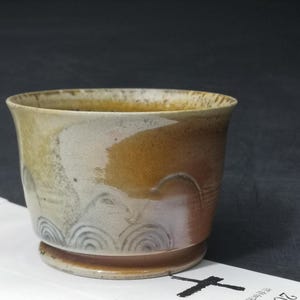 May include: A ceramic cup with a wide mouth and a rounded base. The cup features a blend of brown, tan, and white hues, with a decorative swirl pattern near the bottom. The cup sits on a white paper with Japanese text and the date 2022.10.17.
