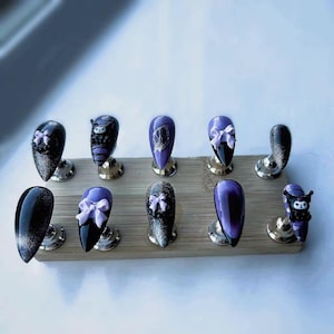 May include: A collection of ten artificial nails in various designs. The nails are displayed on a wooden base. Designs include black and silver glitter, purple, and black with white bows. Some nails feature character art.