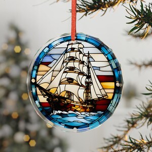 May include: Round Christmas ornament with a stained glass design of a tall ship sailing on blue waves. The ornament has a red ribbon for hanging and is surrounded by a blue border. The ship has white sails and a brown hull.