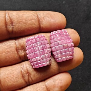 Op de afbeelding: Twee gebeeldhouwde, roze edelstenen met een rasterachtig patroon. De edelstenen worden in een hand tegen een donkere achtergrond gehouden. De stenen hebben een rechthoekige vorm met afgeronde randen.