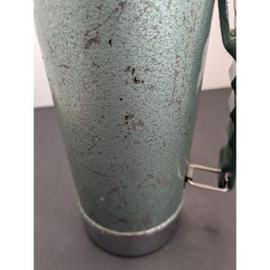 May include: A cylindrical metal thermos with a textured, sage green finish showing signs of wear and scratches. It features a dark green plastic handle attached with silver metal hardware and a smooth, polished silver metal base at the bottom.