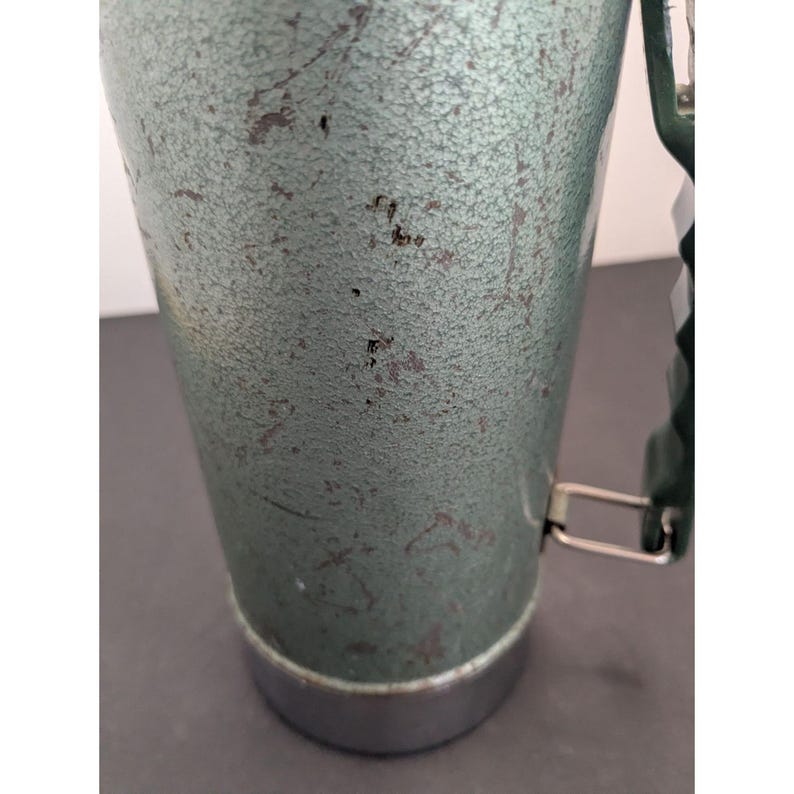 May include: A cylindrical metal thermos with a textured, sage green finish showing signs of wear and scratches. It features a dark green plastic handle attached with silver metal hardware and a smooth, polished silver metal base at the bottom.