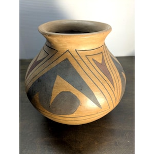 May include: A tan-colored ceramic pot with a rounded body and a slightly flared rim. The pot is decorated with geometric patterns in black, gray, and brown. The design features triangles and curved shapes. The pot has a dark interior.