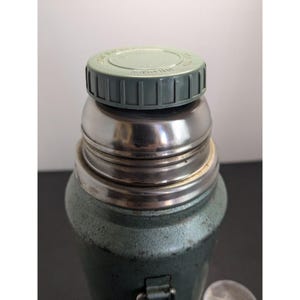 May include: A vintage-style green metal thermos featuring a textured finish, a polished stainless steel neck, and a light green plastic screw-on cap with embossed text on the top surface. The container has a cylindrical body and a sturdy metal handle.