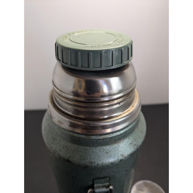 May include: A vintage-style green metal thermos featuring a textured finish, a polished stainless steel neck, and a light green plastic screw-on cap with embossed text on the top surface. The container has a cylindrical body and a sturdy metal handle.