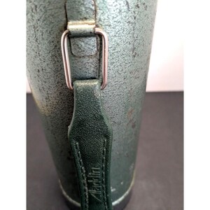 May include: A cylindrical metal thermos with a textured, sage green finish. It features a dark green vertical handle attached by a silver-toned rectangular metal loop. The word Aladdin is embossed vertically into the center of the handle strap.