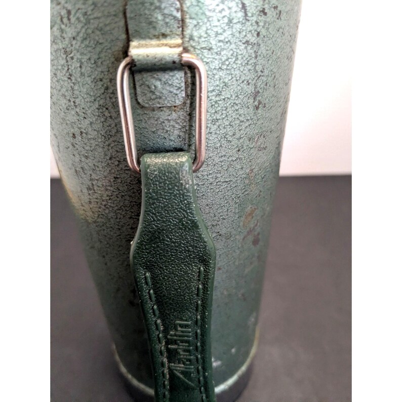 May include: A cylindrical metal thermos with a textured, sage green finish. It features a dark green vertical handle attached by a silver-toned rectangular metal loop. The word Aladdin is embossed vertically into the center of the handle strap.