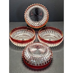 May include: Four clear glass bowls with a red rim and diamond pattern. The bowls have a starburst design in the center and a sawtooth edge. The bowls are arranged on a dark surface.