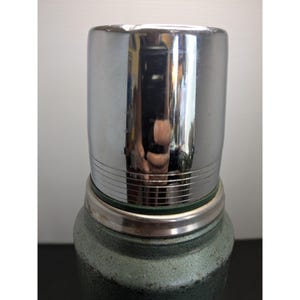 May include: A vintage-style vacuum insulated thermos featuring a textured, dark green hammered metal finish on the body and a polished, reflective chrome-plated metal cup lid with horizontal grooved detailing around the base of the cap.