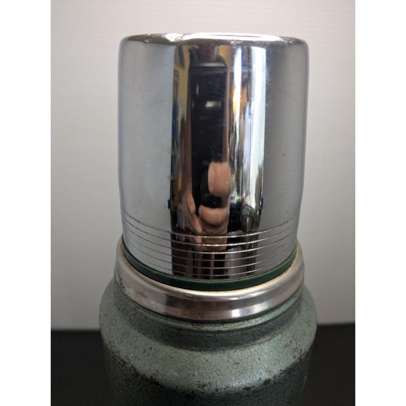 May include: A vintage-style vacuum insulated thermos featuring a textured, dark green hammered metal finish on the body and a polished, reflective chrome-plated metal cup lid with horizontal grooved detailing around the base of the cap.
