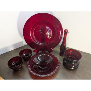 May include: A collection of ruby red glassware, including a large plate, small bowls, a decorative bottle, and a decorative bowl. The plate is displayed on a wooden stand. The glassware is arranged on a dark surface.