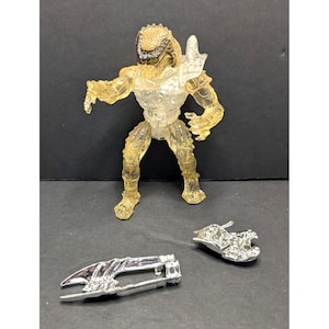 May include: A translucent action figure of a Predator alien with a yellow-tinted body and silver accessories. The figure is posed in a dynamic stance, with detailed armor and weaponry. Additional silver accessories are included.