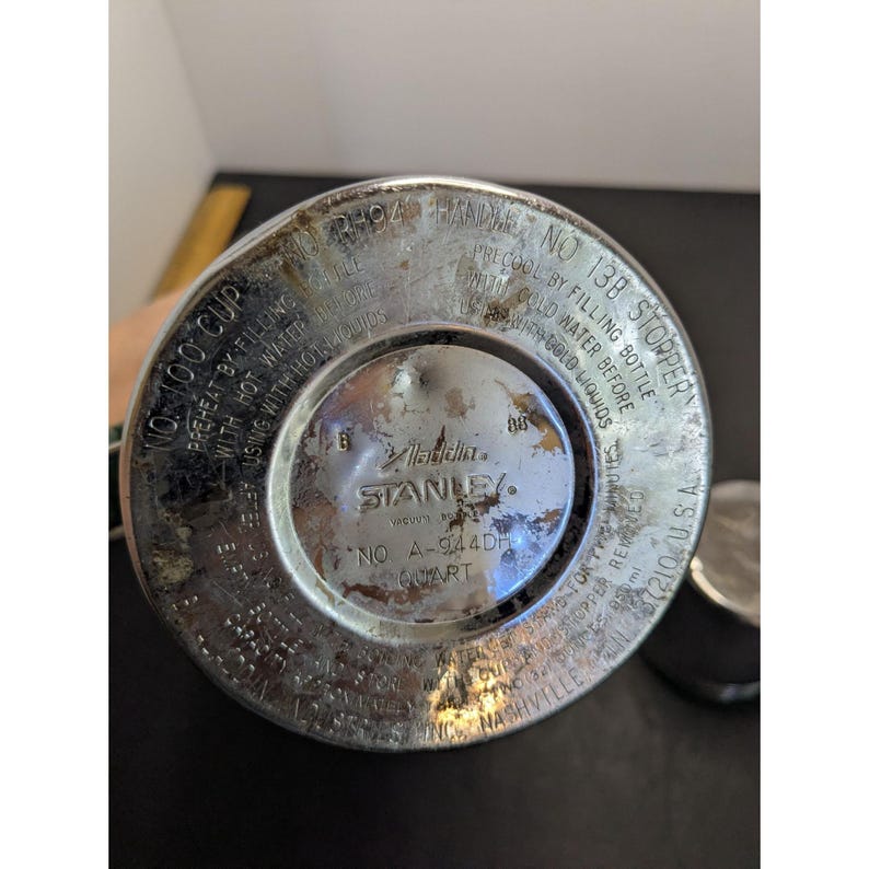 May include: The circular metal base of a silver Stanley Aladdin vacuum bottle. The surface features engraved text including 'NO 100 CUP', 'NO 13B STOPPER', 'NO A-944DH', 'QUART', and 'STANLEY'. The metal shows signs of wear, oxidation, and discoloration.