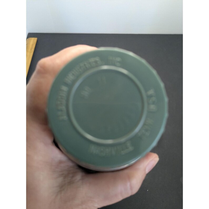 May include: A circular, olive green lid for a container featuring embossed text that reads ALADDIN INDUSTRIES INC, NASHVILLE TENN USA, and NO 19. The lid has a raised outer rim and a recessed center section, with a smooth, matte finish throughout.