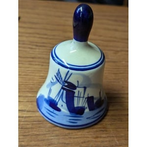 May include: A ceramic bell with a blue handle and a white body. The bell features a blue windmill design and blue accents. The bell is likely a decorative item or souvenir.