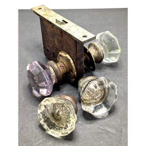 May include: A vintage mortise lock mechanism featuring a rectangular rusted metal case and three faceted glass doorknobs. One knob is attached to the lock, while two others are separate. The knobs have clear and light purple glass surfaces.