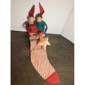 May include: A Christmas stocking with a red toe and a pink and white striped body. Two elf dolls with red hats and green and red clothing are at the top. A pink star is attached to the stocking.