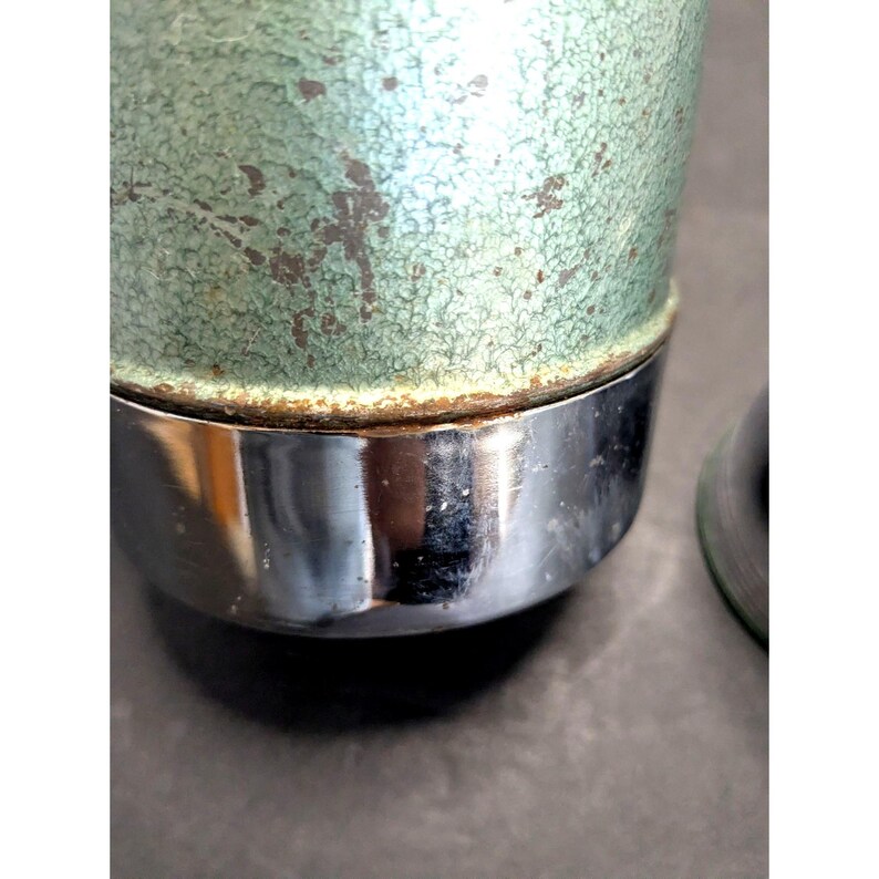 May include: A cylindrical container featuring a textured, mint green crackle finish on the upper portion and a smooth, reflective chrome base. The junction between the green and chrome sections shows visible signs of oxidation and rust.