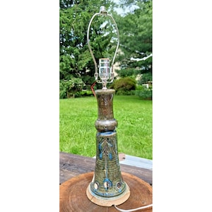 May include: A tall, ceramic table lamp with a green and brown glazed finish. The lamp features a decorative design with blue accents and a silver-toned metal lamp holder. The lamp sits on a round, natural wood base.