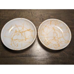 May include: Two white ceramic bowls with a gold, abstract, web-like design. The bowls are round with a shallow depth, and the gold lines create a unique pattern across the surface. The bowls are placed on a dark wooden surface.