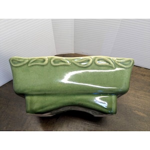 May include: A rectangular, green ceramic planter with a glossy finish. The top edge features a repeating leaf design. The planter has a curved base and is suitable for indoor or outdoor use. The planter is approximately 10 inches long.