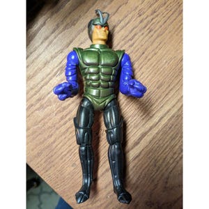 May include: A vintage action figure of a humanoid insect-like character. The figure has a green torso, blue arms, black legs, and a silver helmet. The eyes are red, and the figure is posed with its fists clenched.