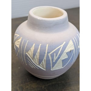 May include: A small, round, ceramic vase with a light purple exterior and a cream-colored interior. The vase features a geometric design in a lighter color, possibly white or silver. The vase has a wide opening and a short neck.