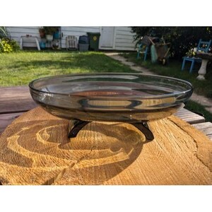 May include: A clear, shallow glass bowl with a dark gray tint, resting on a black metal stand. The bowl is circular and sits on a wooden surface. The bowl is approximately 12 inches in diameter.