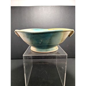 May include: A ceramic bowl featuring a wide, shallow basin with a pedestal base and two small, integrated side handles. The exterior surface displays a blended glaze of deep teal, turquoise, and soft beige tones with a smooth, curved silhouette.